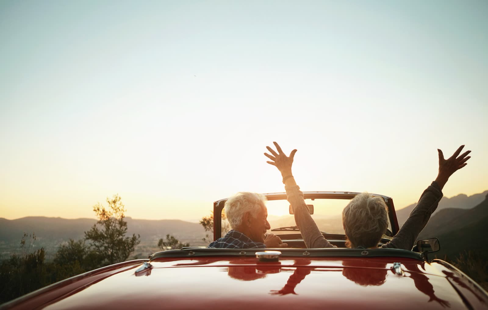 Older couple in a red convertible raising their arms toward a mountain sunset